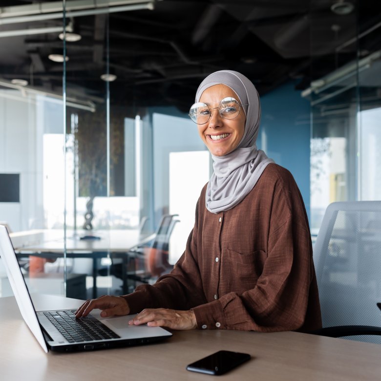 Kvinne med hijab og briller jobber på bærbar datamaskin i et moderne kontor.