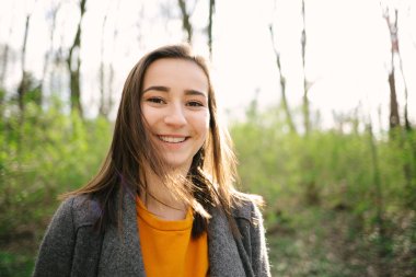 Kvinne smiler utendørs i en skog med sollys i bakgrunnen.