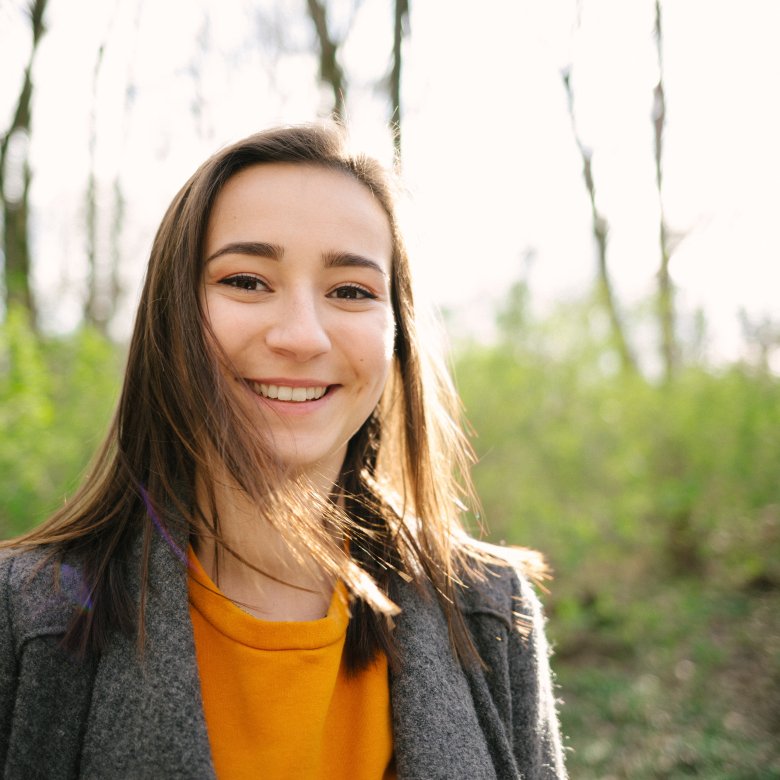 Kvinne smiler utendørs i en skog med sollys i bakgrunnen.