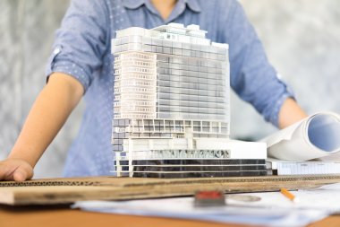 Person holder en modell av en moderne bygning med arkitekttegninger på bordet.