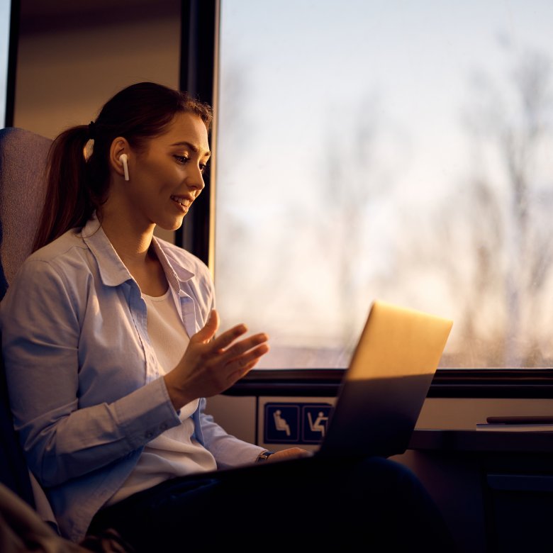 Kvinne med laptop og ørepropper smiler og gestikulerer på et tog.