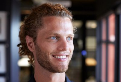 Mann med krøllete hår og skjegg smiler og ser til siden i et kontormiljø.