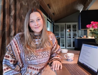 Kvinne i strikkegenser studerer på laptop ved et trebord, med en kopp te ved siden av.