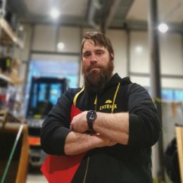 Mann med skjegg og hettegenser står med armene i kors i et verksted.