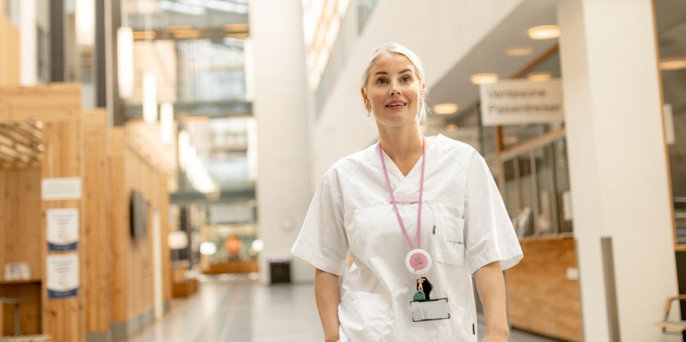 Sykepleier i hvit uniform står i en moderne sykehusgang.