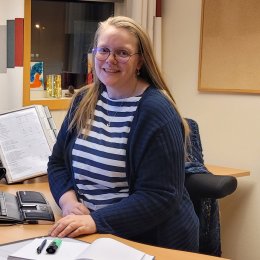 Kvinne smiler ved skrivebord med datamaskin, notatbok og vannflaske.