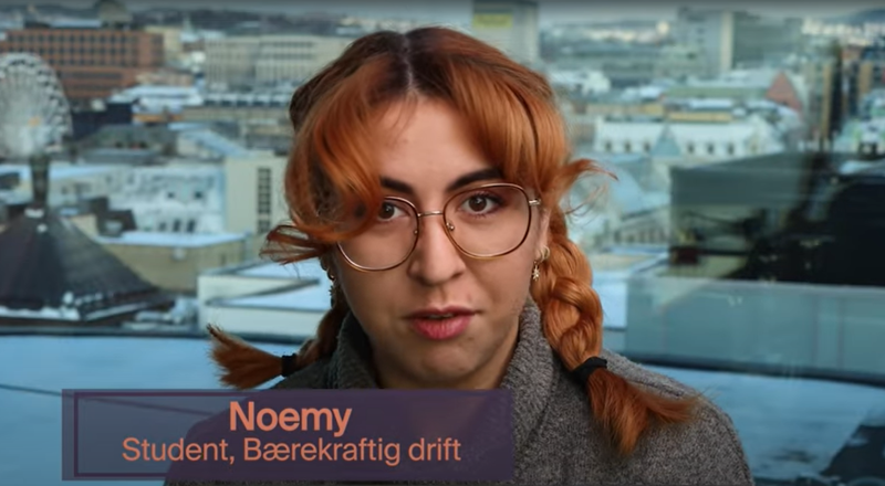 Student med briller og fletter snakker om bærekraftig drift foran bylandskap.