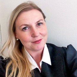 Kvinne med langt blondt hår og sort kappe smiler foran en hvit vegg.