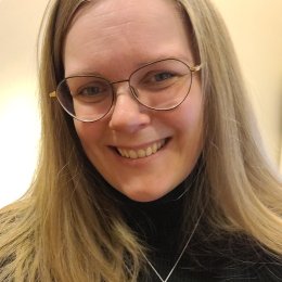 Kvinne med langt blondt hår og briller smiler mot kameraet.