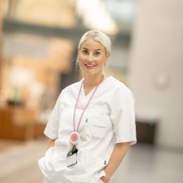 Kvinne i hvit uniform med rosa stetoskop smiler i en lys, moderne bygning.