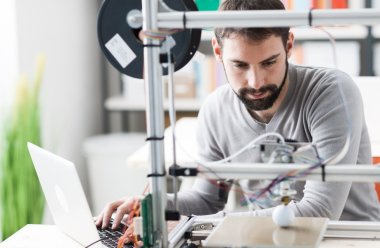 Mann jobber med 3D-printer og bærbar datamaskin i et kontormiljø.