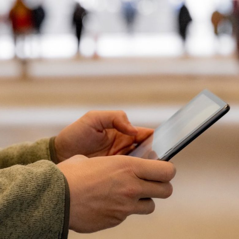 Person holder en mobiltelefon, uskarpe mennesker i bakgrunnen.