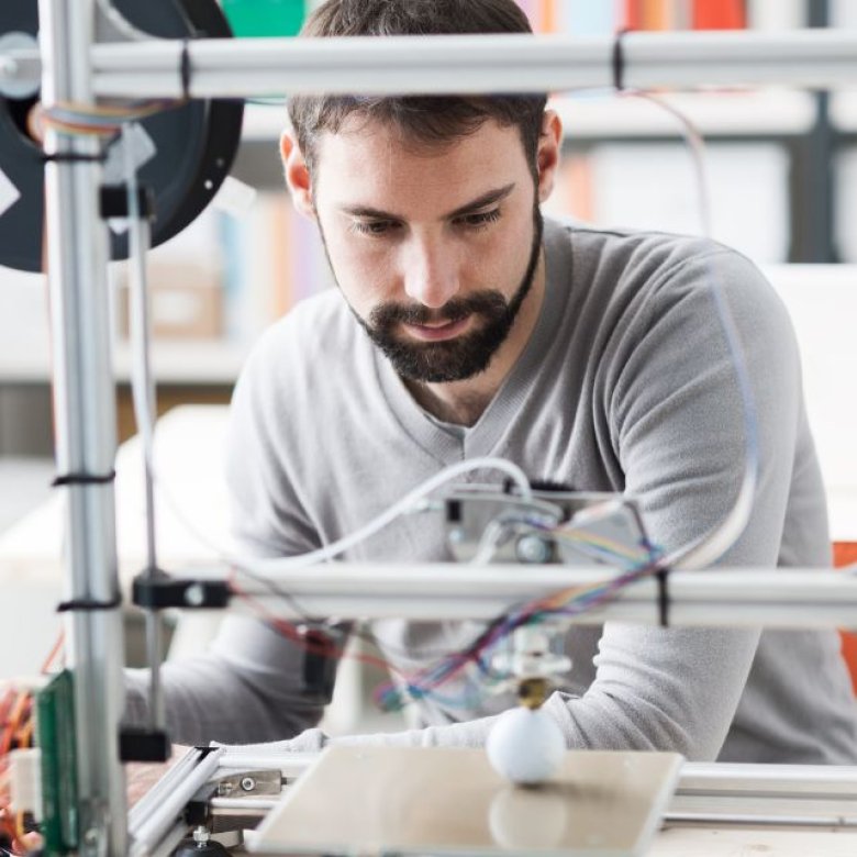 Mann jobber med 3D-printer og bærbar datamaskin i et kontormiljø.