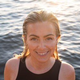 Kvinne smiler foran vannet i solnedgang, iført svart badedrakt.