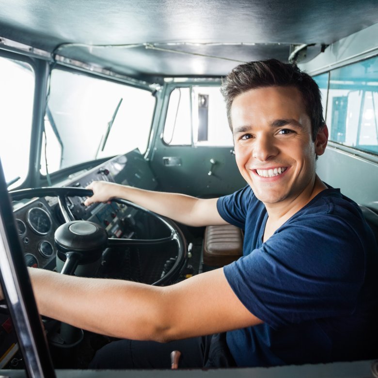 Smilende mann sitter bak rattet i en buss eller lastebil, klar til å kjøre.