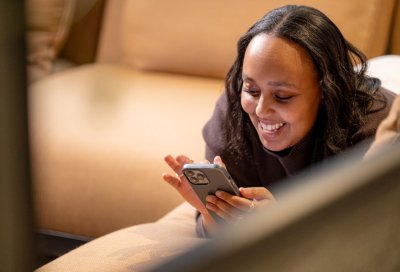 Kvinne smiler mens hun ser på mobilen, liggende på en sofa.