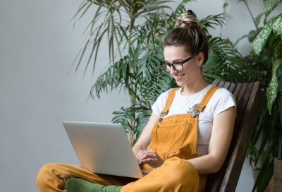 Kvinne i gule snekkerbukser jobber på laptop, omgitt av grønne planter.