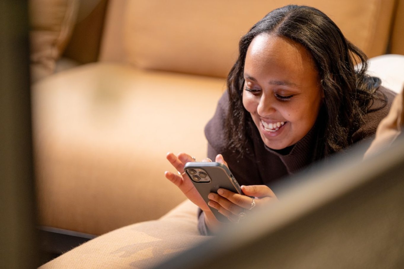 Kvinne smiler mens hun ser på mobilen, liggende på en sofa.