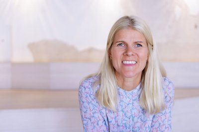 Kvinne med blondt hår smiler iført mønstret topp, sittende foran en lys bakgrunn.