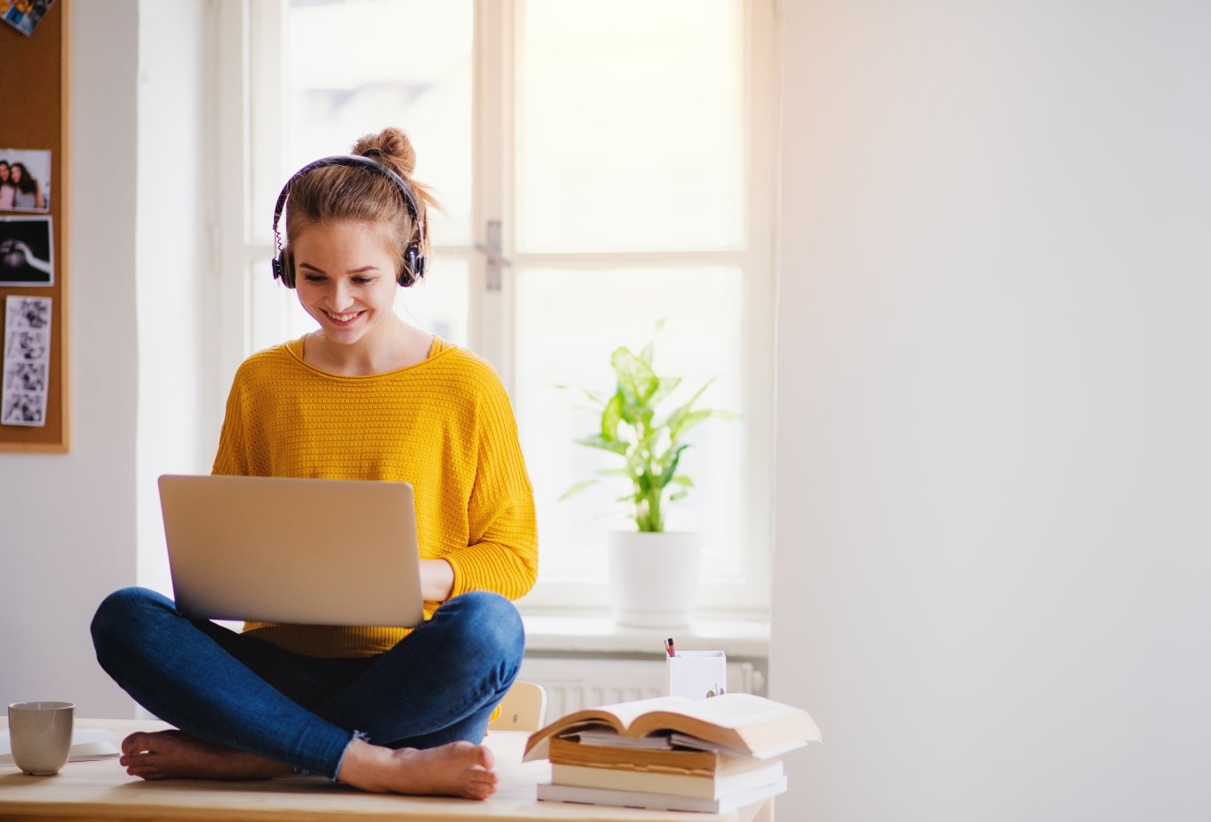 Student med hodetelefoner jobber på laptop ved vindu, med bøker og plante i bakgrunnen.