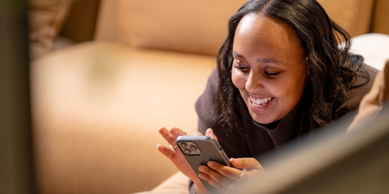 Kvinne smiler mens hun ser på mobilen sin, liggende på en sofa.