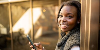 Ung kvinne smiler og holder en mobiltelefon utendørs.