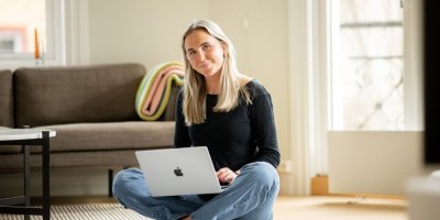 Kvinne sitter på gulvet med en laptop i fanget, smilende i et lyst rom.