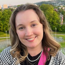 Kvinne smiler utendørs med grønn park og bylandskap i bakgrunnen.