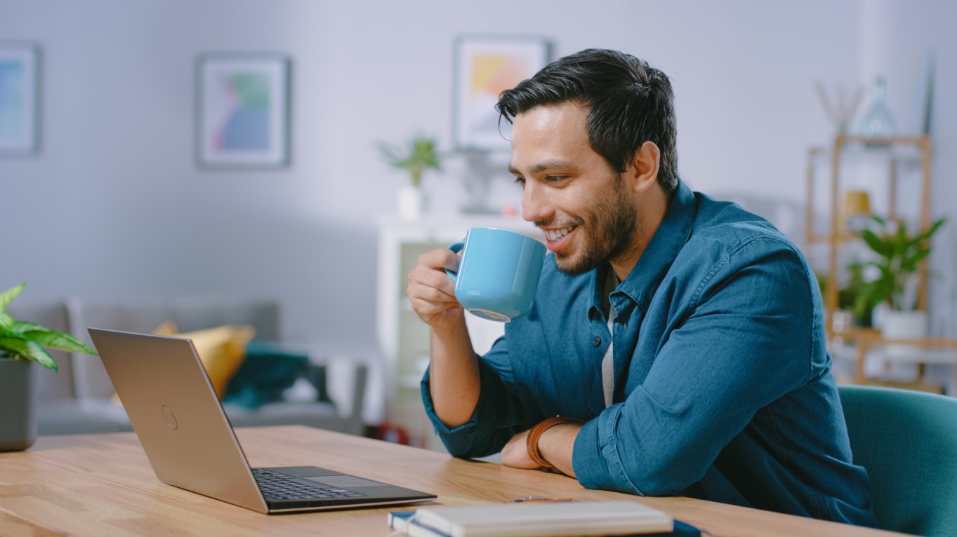 mann smiler og drikker kaffe og ser på pc skjerm