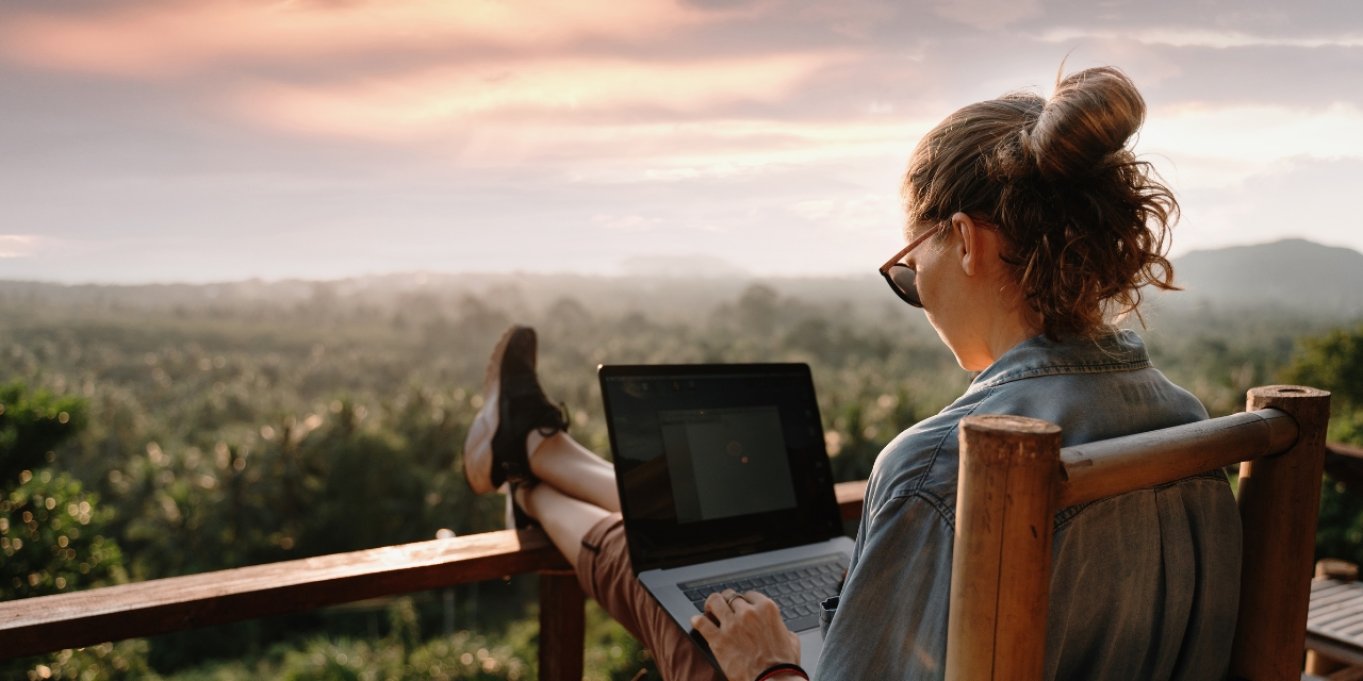 Person som jobber på laptop med utsikt over skog og solnedgang.