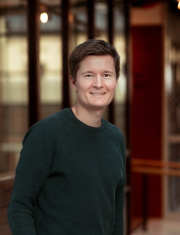 Mann i grønn genser smiler foran en moderne kontorbygning.