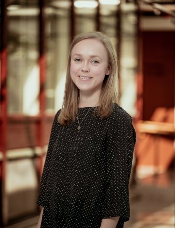 Kvinne med langt hår og svart kjole smiler foran en glassvegg.