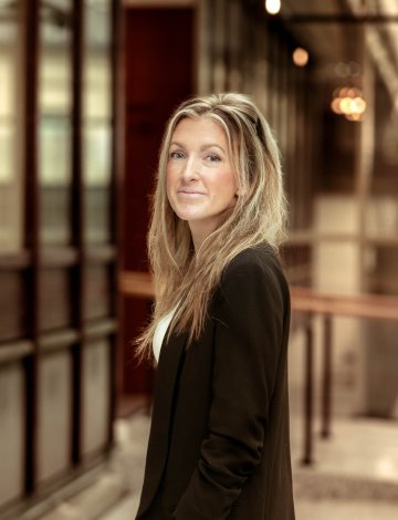 Kvinne med langt hår og svart jakke smiler i en kontorbygning.