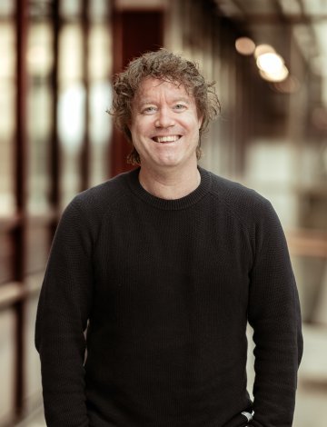 Mann med krøllete hår og svart genser smiler i en gang med glassvegger.