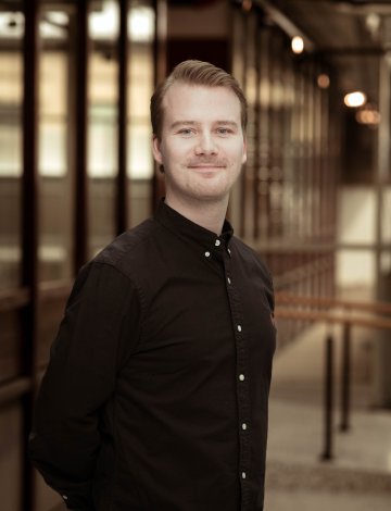 Mann i svart skjorte smiler foran en glassvegg i en kontorbygning.