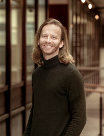 Mann med langt hår og svart genser smiler foran en bygning med glassvinduer.