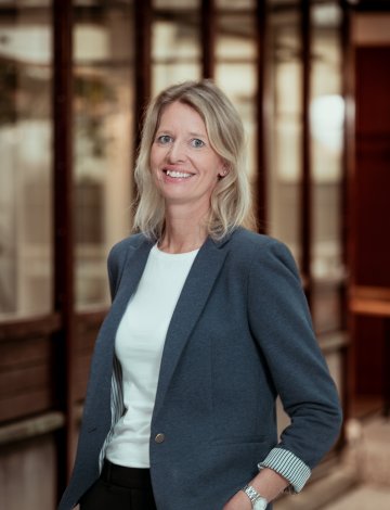 Kvinne i blå blazer og hvit topp smiler foran en glassvegg.