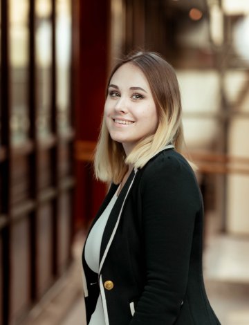 Kvinne med blondt hår og svart jakke smiler foran en glassvegg.