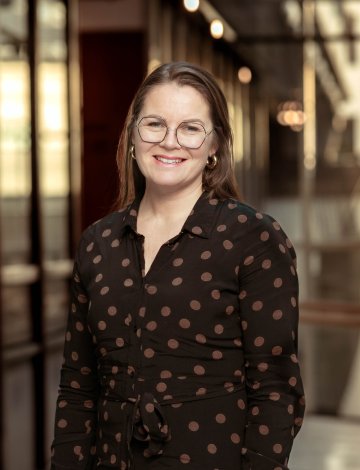 Kvinne med briller og prikkete skjorte smiler foran en glassvegg.