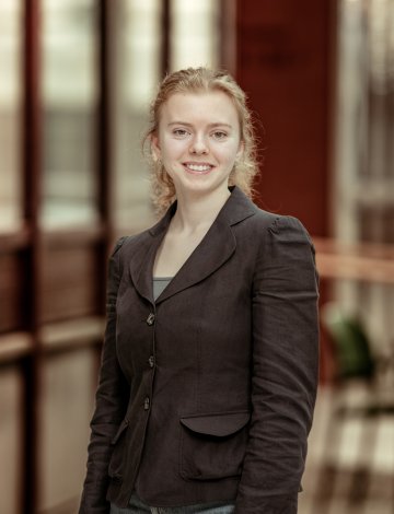 Kvinne i svart jakke smiler foran en glassvegg i en kontorbygning.