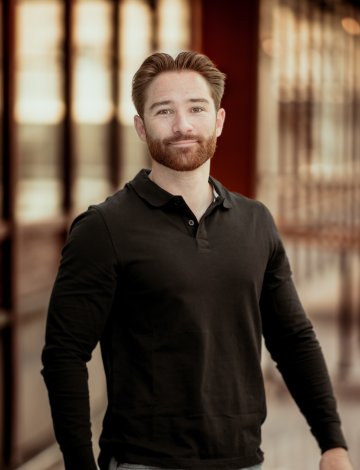 Mann med skjegg og svart genser smiler foran en glassvegg.