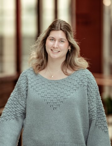 Kvinne med langt hår og grå genser smiler foran en uskarp bakgrunn.