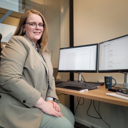 Kvinne i dress sitter ved skrivebord med to dataskjermer på kontor.