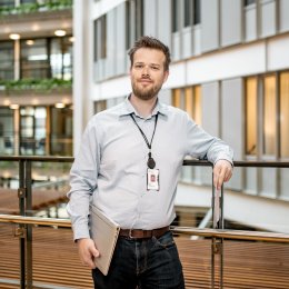 Mann med skjegg og ID-kort står i moderne kontorbygg, holder en bærbar datamaskin.