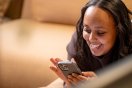 Kvinne smiler mens hun ser på mobilen sin, liggende på en sofa.