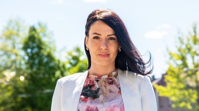 Kvinne i lys jakke og blomstrete topp smiler utendørs på en solrik dag.