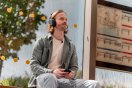Mann med hodetelefoner sitter på en benk ved et busstopp, holder en mobiltelefon.