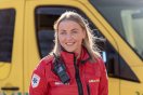 Ambulansearbeider i rød uniform foran en gul ambulanse, smilende og klar til innsats.