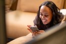 Kvinne smiler mens hun ser på mobilen, liggende på en sofa.