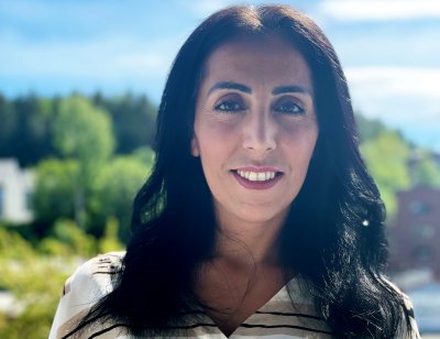Kvinne smiler foran et grønt landskap med trær og bygninger i bakgrunnen.